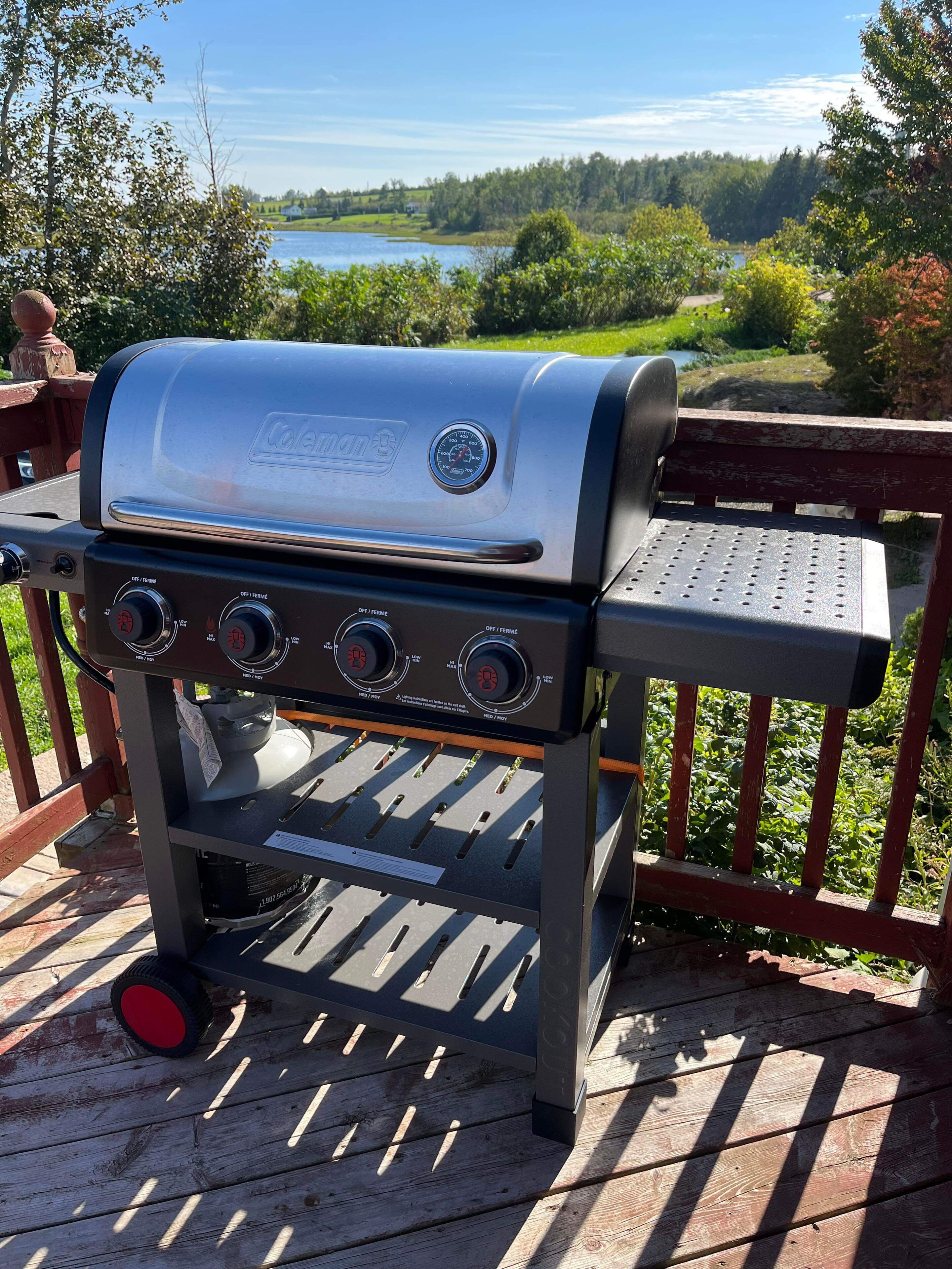 A Coleman gas grill on an outdoor deck with a view of a lake and forest.