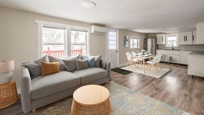 Una sala de estar moderna con un sofá gris, una mesa de centro redonda de madera y un comedor con mesa y sillas blancas.