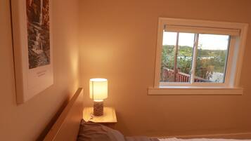 A bedroom with a bed, a nightstand with a lamp, a window with a view, and a framed picture on the wall.