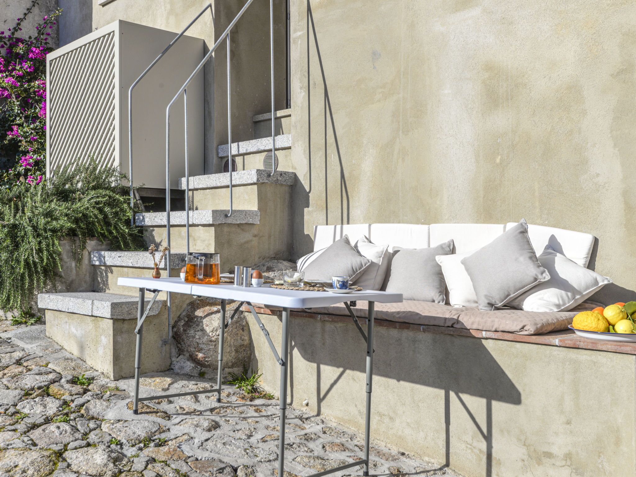 Une terrasse en pierre avec une table et des chaises en métal, un canapé blanc avec des coussins et une assiette de citrons.