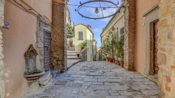 Una estrecha calle empedrada con edificios de piedra, un arco y campanas colgantes.