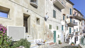Una estrecha calle empedrada flanqueada por edificios con balcones, ventanas con contraventanas y una pequeña mesa con objetos encima.