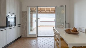 Een moderne keuken met een inbouwoven, een eettafel met stoelen en uitzicht op zee door een open deur.