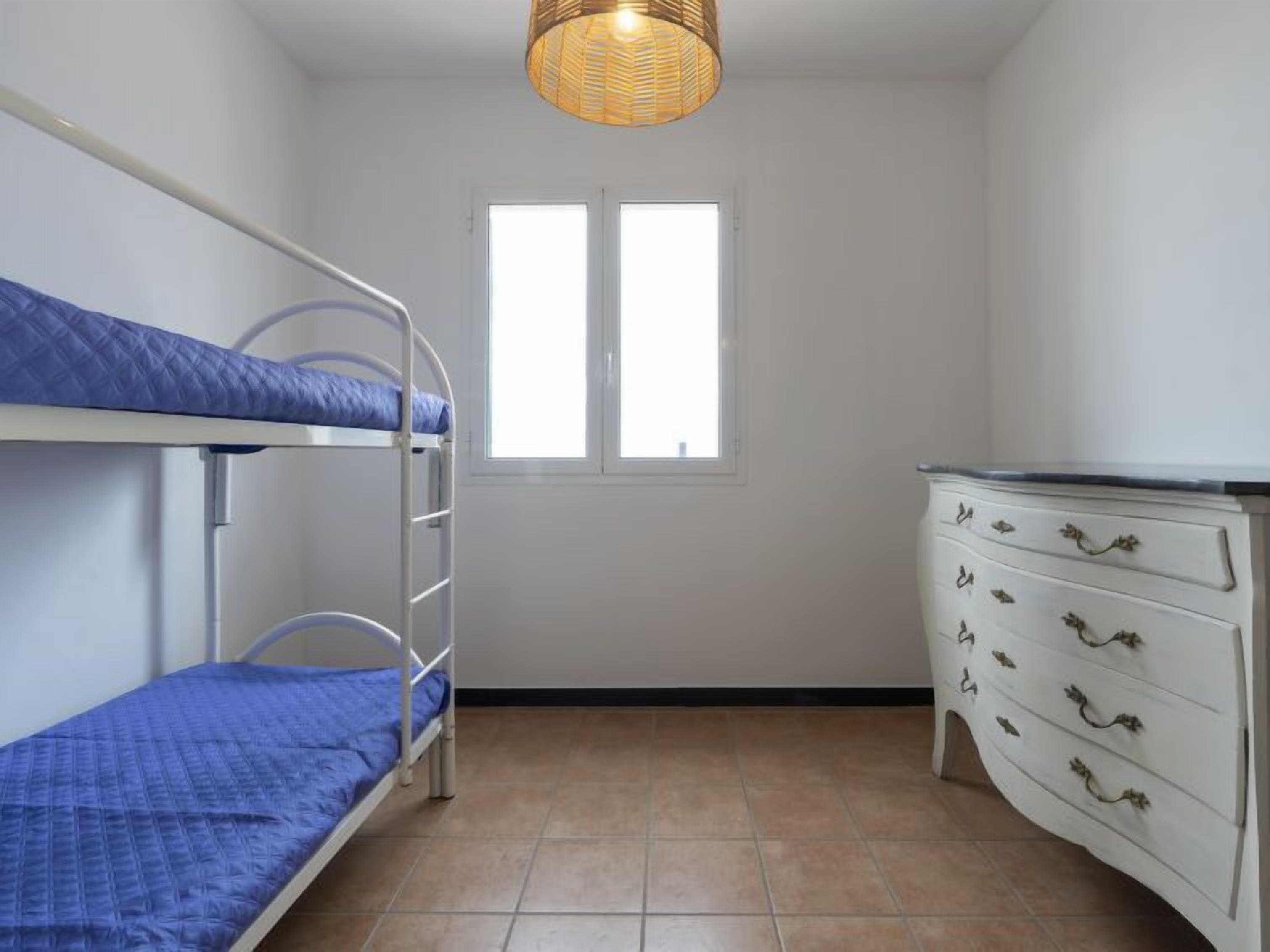 Une petite pièce bien éclairée, avec des lits superposés, une commode et une fenêtre.