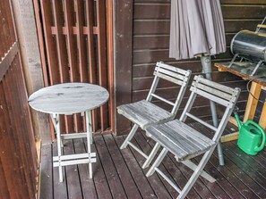En träveranda med ett runt bord och två stolar, en grön vattenkanna och ett grått parasoll.