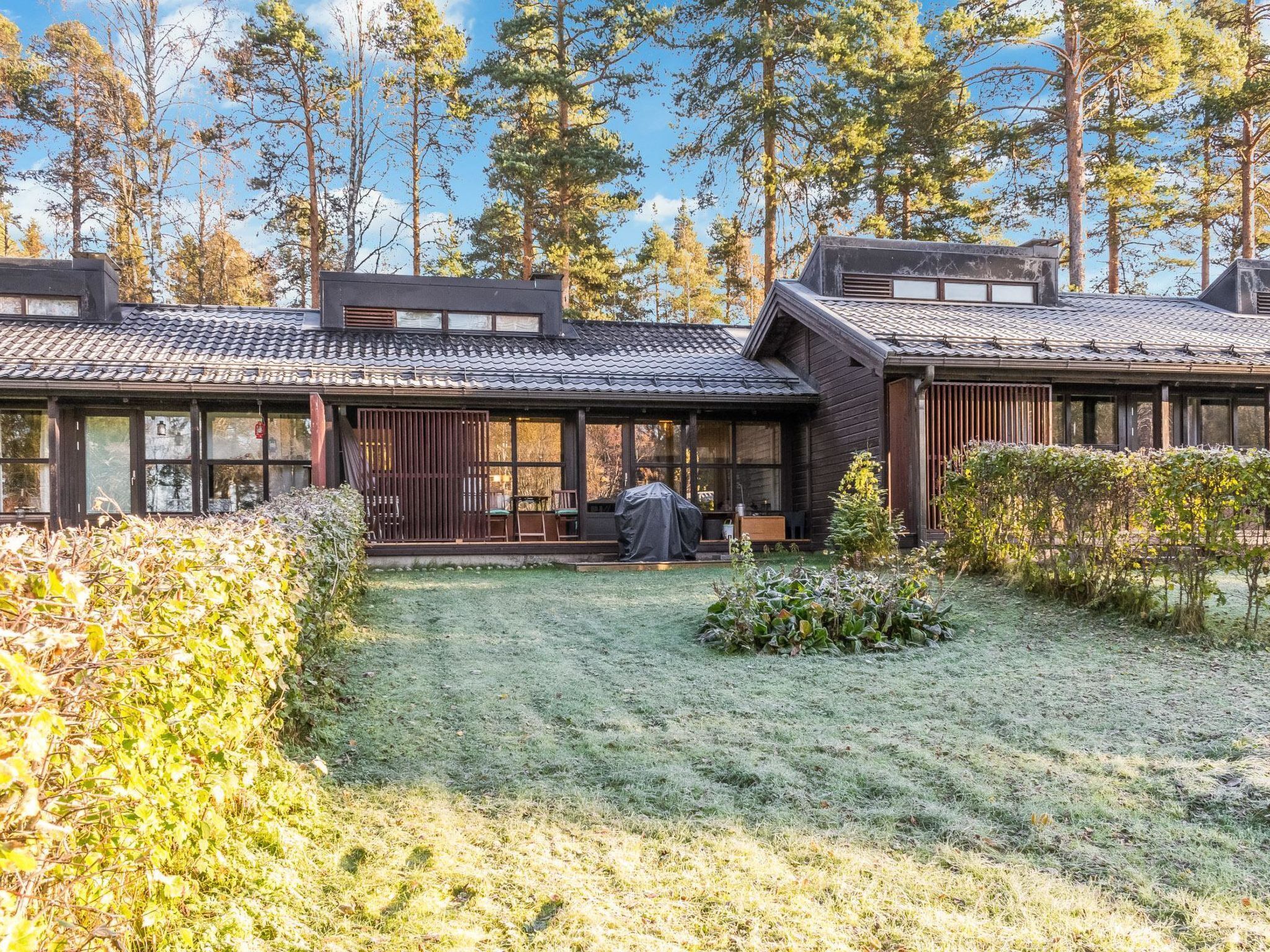 Perinteinen puutalo, jossa on katettu terassi, ja jonka ympärillä on nurmikko ja korkeita puita.