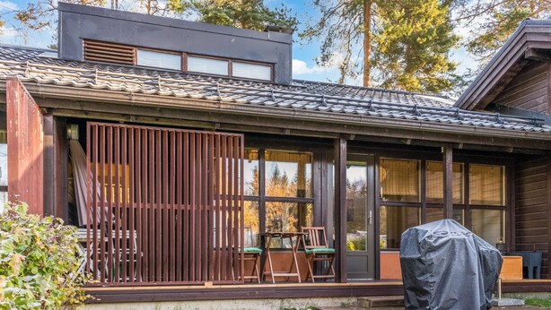 A modern house with a covered porch, a grill, and a view of the surrounding trees.