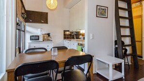 A compact kitchen with white cabinets, a microwave, and a coffee maker. There is a dining table with chairs and a small white cart. A ladder is leaning against the wall.
