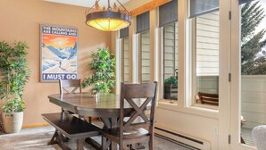 A dining area with a wooden table and chairs, a poster on the wall, and a large window with blinds. - SunVail Condo - Steps from Bridge to Lionshead (Vail)