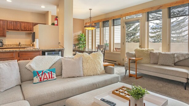A modern living room with a sofa, a coffee table, and a fireplace. There is a kitchen area with wooden cabinets and a dining table. - SunVail Condo - Steps from Bridge to Lionshead (Vail)