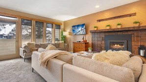 A cozy living room with a fireplace, a sofa set, a coffee table, and a large window overlooking a snowy landscape. - SunVail Condo - Steps from Bridge to Lionshead (Vail)