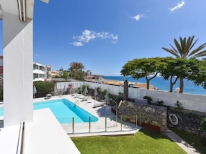 Zona de piscina con vista al mar, rodeada por una valla blanca y vegetación.