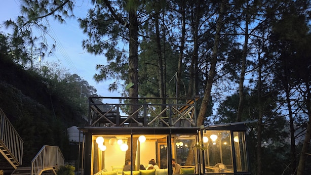 Une cabane aux murs de verre, dotée d’un balcon et entourée d’arbres.