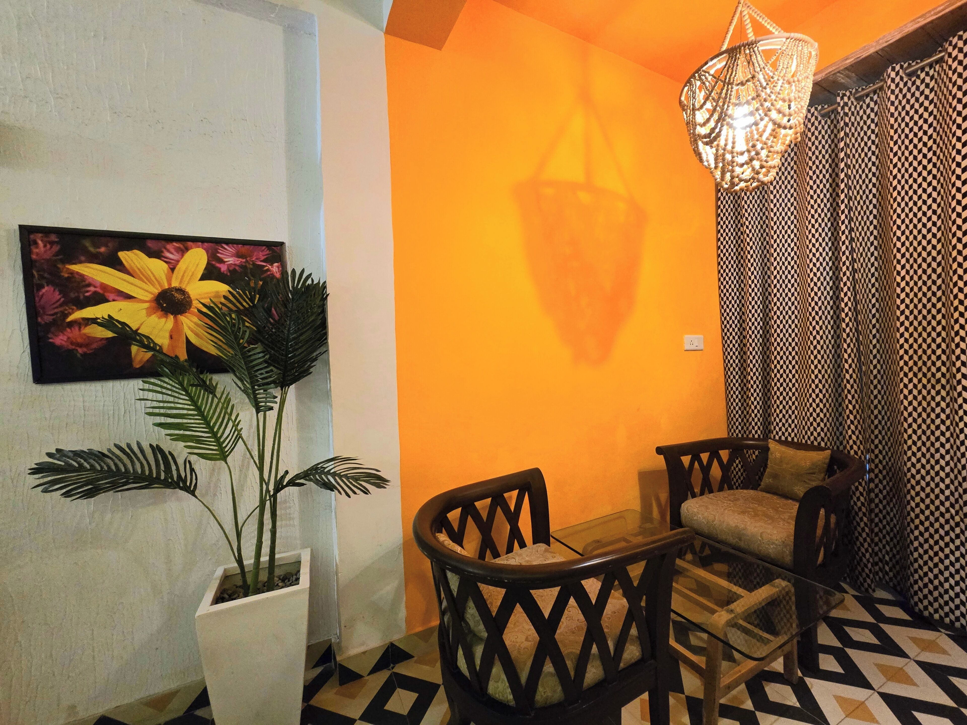 A room with a geometric patterned floor, a white wall with a sunflower painting, a potted plant, a wooden chair, a glass table, and a patterned curtain.