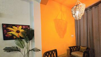 A room with a geometric patterned floor, a white wall with a sunflower painting, a potted plant, a wooden chair, a glass table, and a patterned curtain.