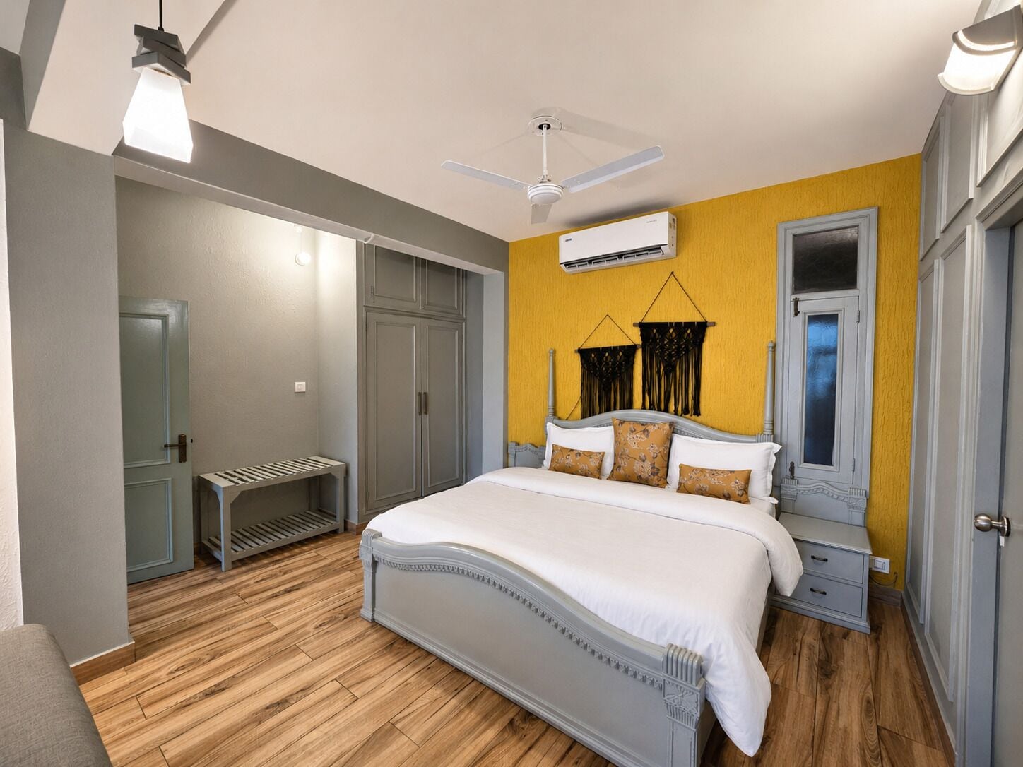 A bedroom with a bed, a wardrobe, a bedside table, a hanging macrame decoration, and an air conditioning unit.