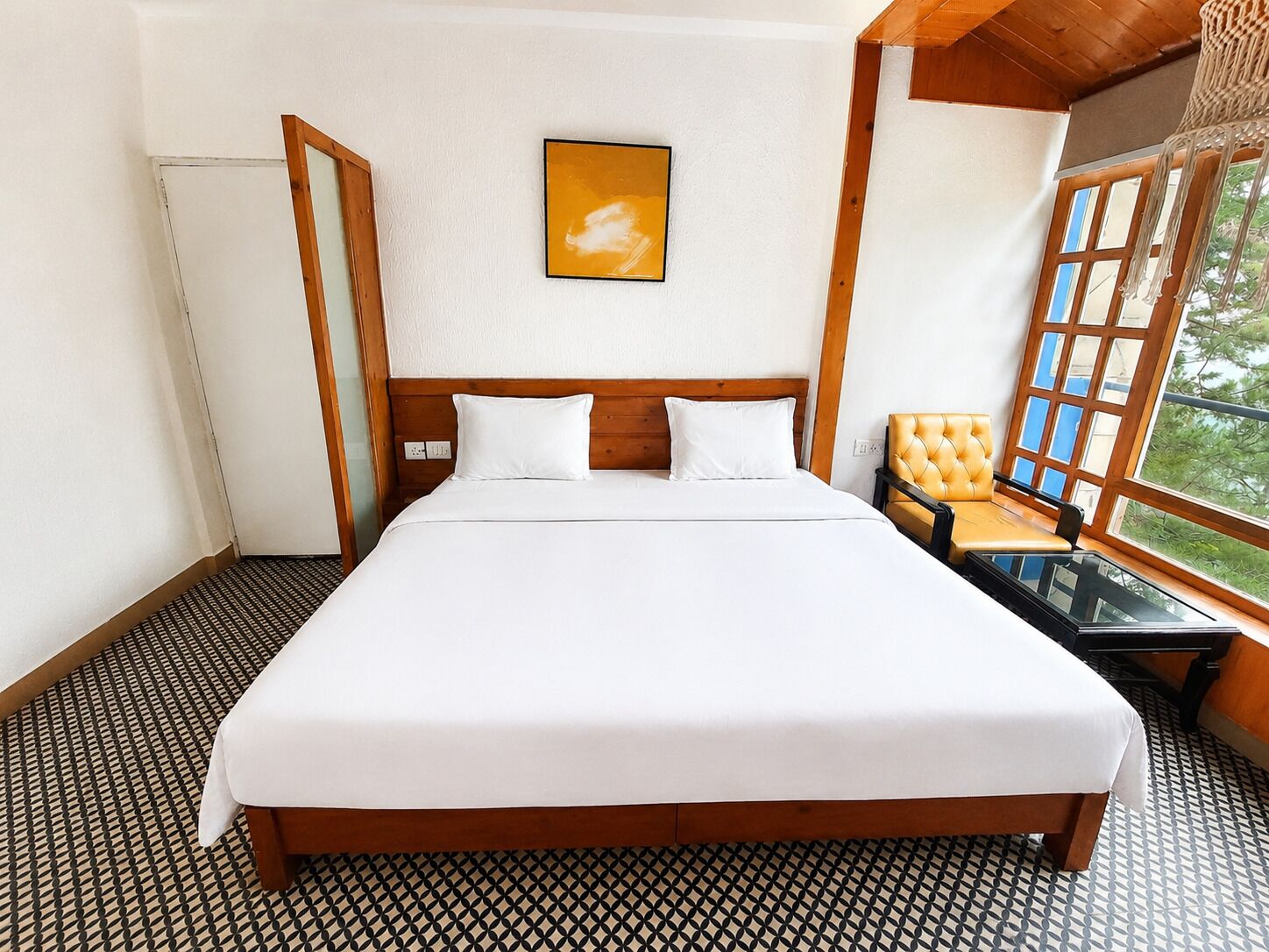 A hotel room with a large bed, a wooden headboard, a single white pillow, a small table with a chair, and a window with a view of greenery.