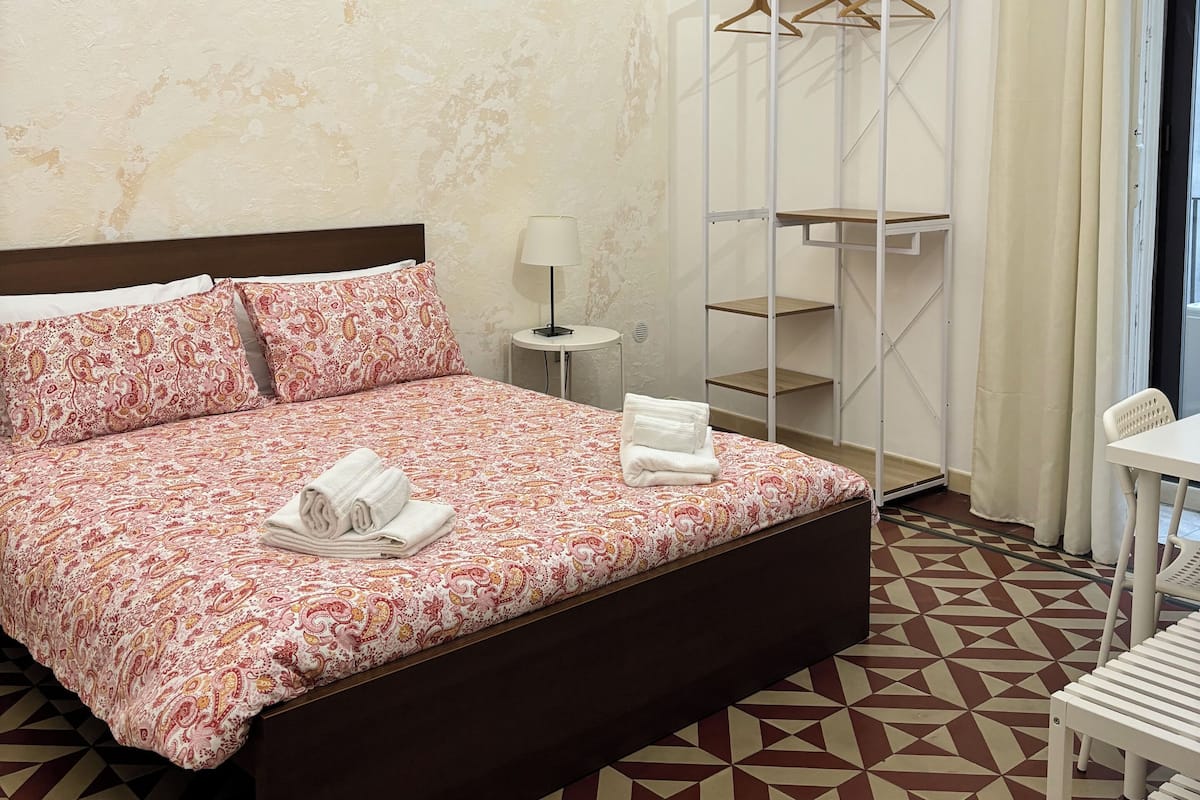 Quarto de hotel com colcha estampada, mesa de cabeceira com abajur, guarda-roupa com cabides e piso com padrão geométrico.