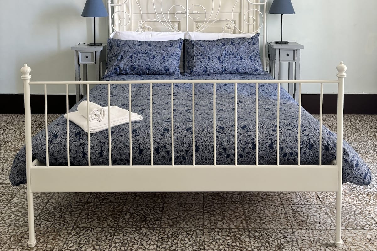 Uma cama com estrutura metálica branca, roupa de cama estampada em azul e duas luminárias de cabeceira.
