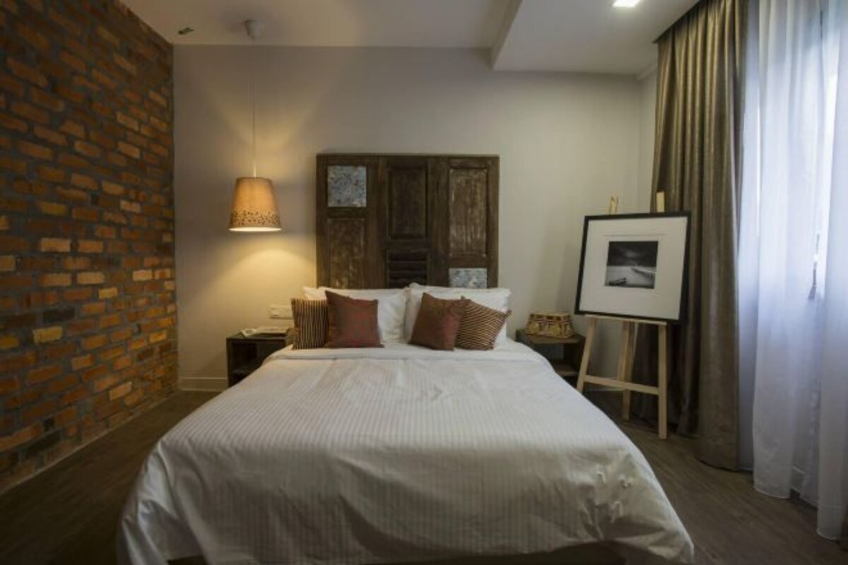 Habitación de hotel con pared de ladrillo, cama con ropa de cama blanca, lámpara colgante, cuadro en la pared y una pequeña mesa con lámpara.