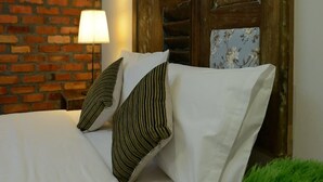 Una cama con sábanas blancas y almohadas a rayas, una pared de ladrillo, un cabecero de madera y una lámpara.