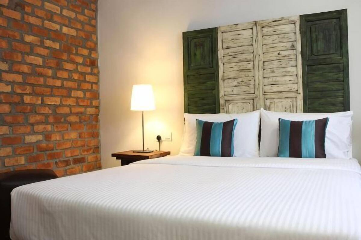A hotel room with a brick wall, a wooden headboard, a bed with white bedding, and a bedside lamp.