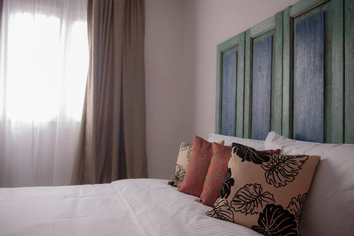A neatly made bed with white linens and decorative pillows, a green-framed headboard, and sheer curtains on a window.