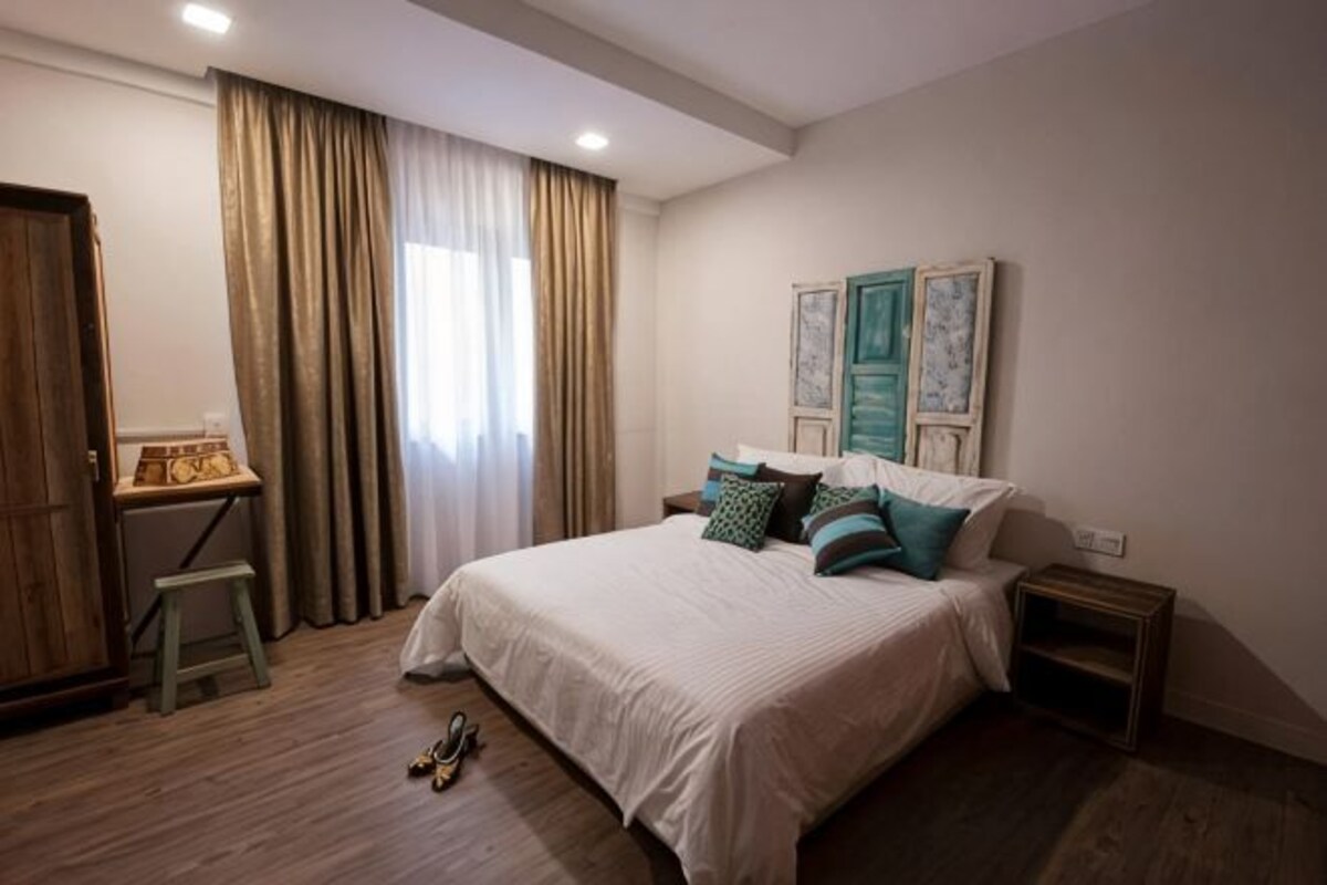 A bedroom with a bed, curtains, a nightstand, a wooden floor, and a small table with a lamp.