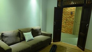 Una sala de estar con sofá, un otomán estampado y una pared de ladrillo visible a través de puertas abiertas.