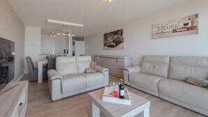 A modern living room with a sofa, a coffee table with a bottle and glasses, and a dining area with a table and chairs. - Modern apartment with beautiful sea view! (Blankenberge)