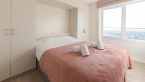 A modern bedroom with a large bed, built-in wardrobe, and a city view through a large window. - Modern apartment with beautiful sea view! (Blankenberge)