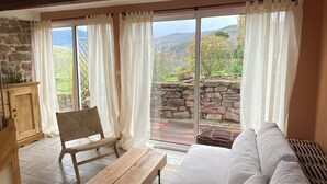 Amplia sala de estar con un banco de madera, un sof á blanco y una chimenea de piedra. Amplias ventanas con cortinas ofrecen vistas del exterior.