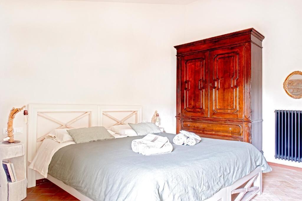 Un letto rifatto con una coperta grigia e cuscini bianchi, un armadio in legno, un termosifone e una piccola mensola con libri e un oggetto decorativo.