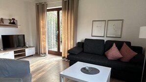Un soggiorno moderno con un divano nero, un tavolino bianco e una televisione su un supporto in legno. È presente un'ampia finestra con tende, e una porta che dà sull'esterno.