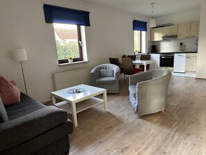 Ein modernes Wohnzimmer mit grauem Sofa, weißem Couchtisch und grauem Sessel. Es gibt einen Essbereich mit Tisch und Stühlen sowie eine Küche mit Schränken und Geräten.