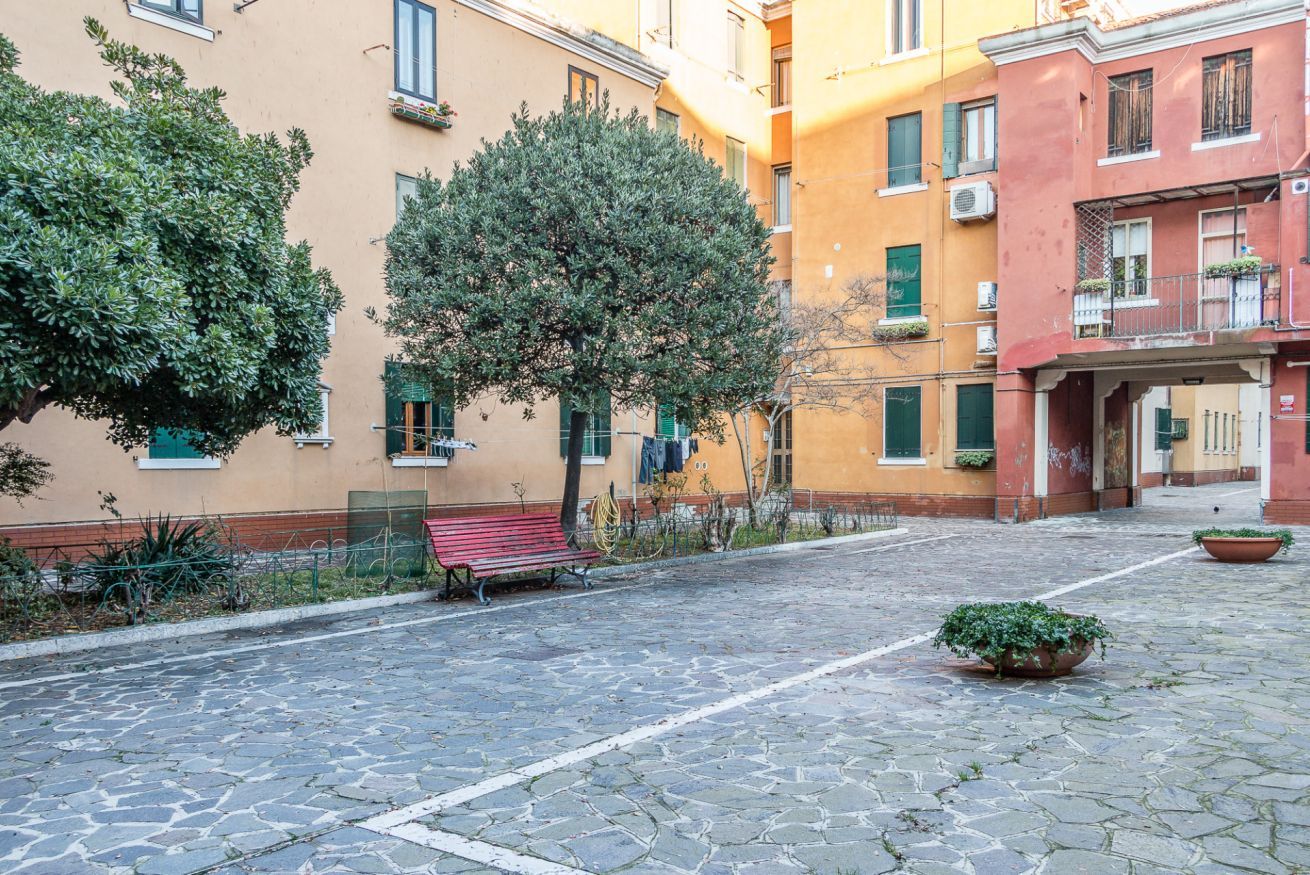 Un patio empedrado con un banco, plantas en macetas y edificios de varios pisos con balcones.