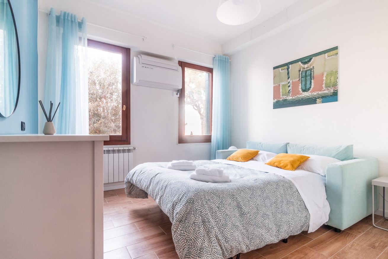 A bedroom with a bed, a bedside table, a window with curtains, an air conditioning unit, and a painting on the wall.