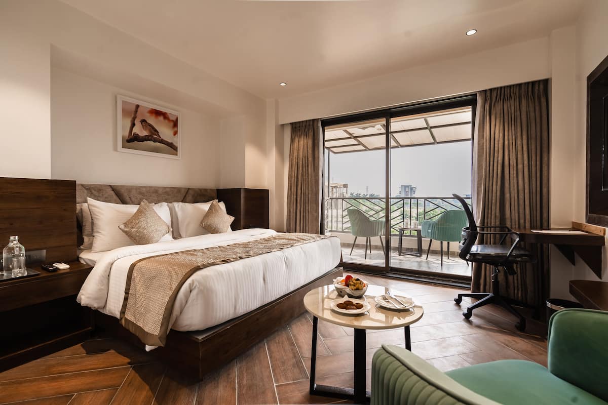 A hotel room with a large bed, a dining table with breakfast, a desk, and a view of the city through the balcony.