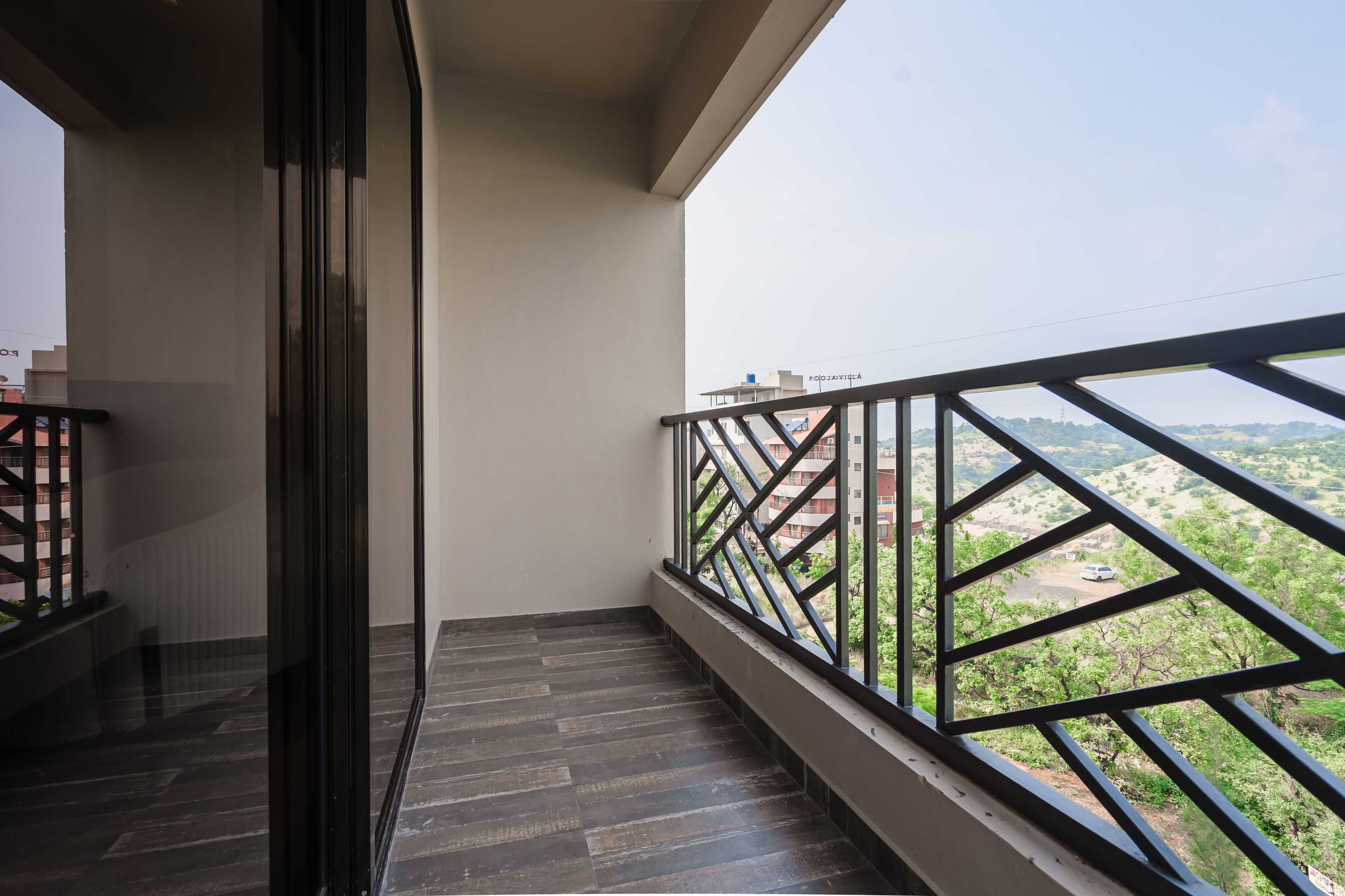 Ein Balkon mit Metallgeländer, Holzboden und Blick auf eine grüne Landschaft mit Gebäuden in der Ferne.