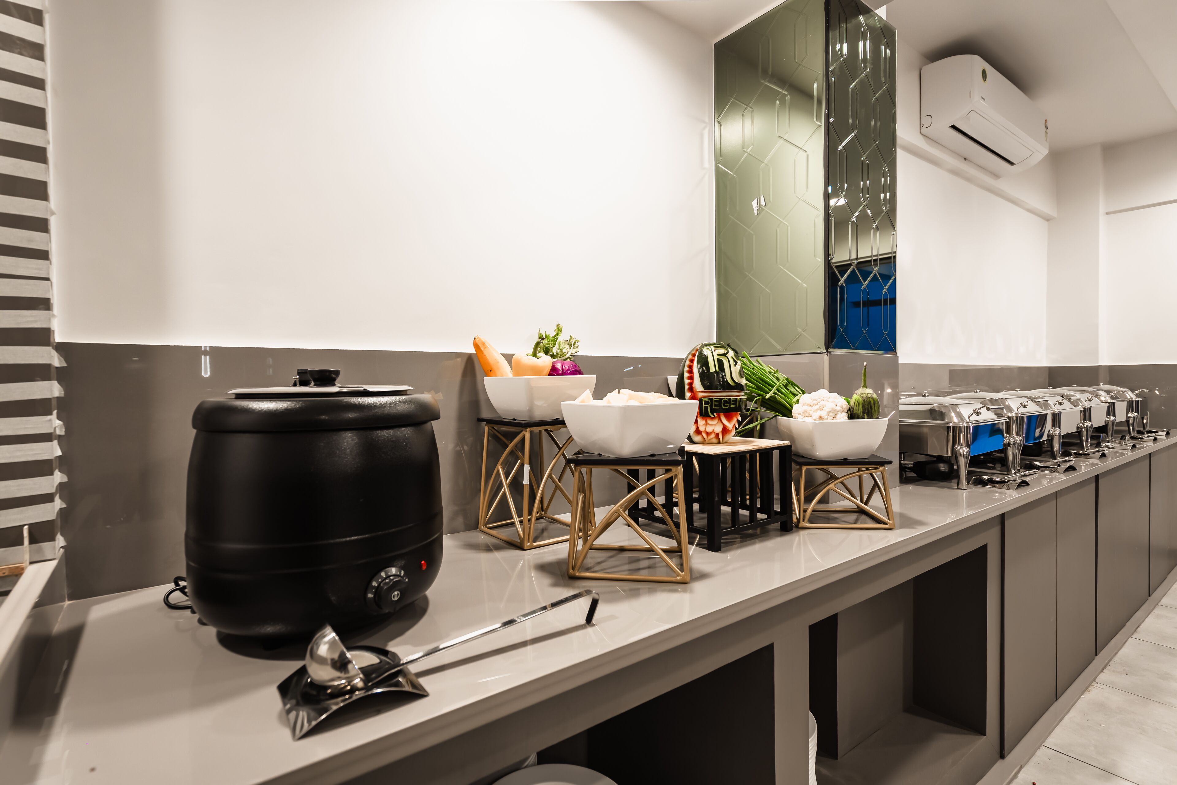 A modern kitchen with a long countertop, a large black water dispenser, and various bowls of fruits and vegetables.
