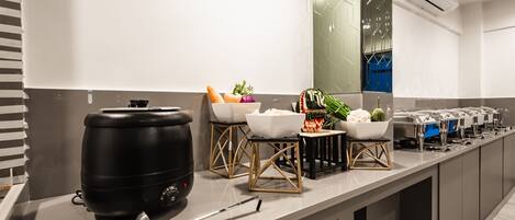 A modern kitchen with a long countertop, a large black water dispenser, and various bowls of fruits and vegetables.