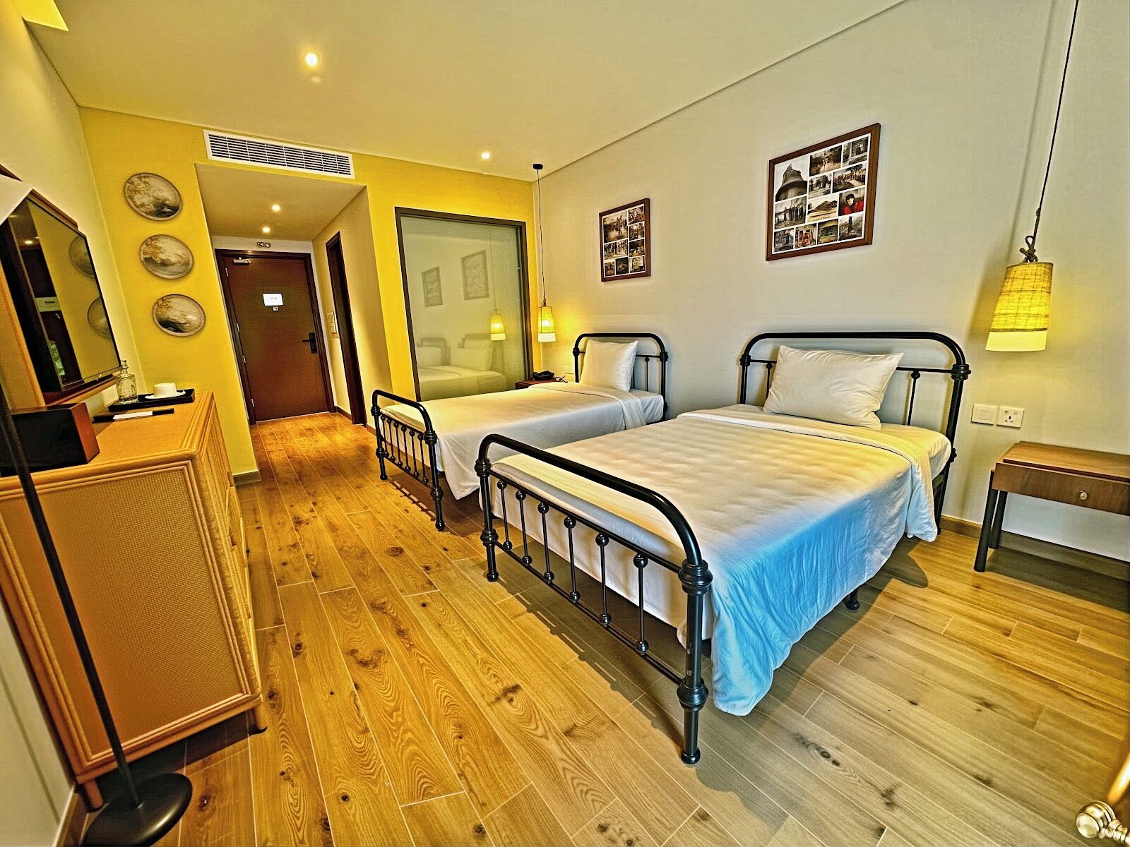 A hotel room with two beds, wooden flooring, a desk, a mirror, and a door leading to another room.