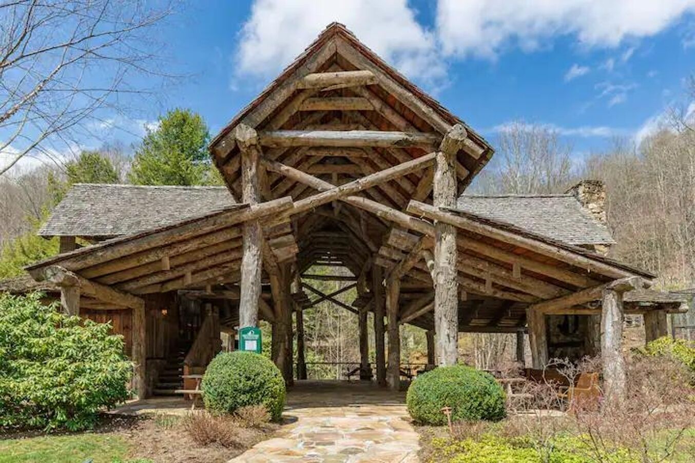 A rustic log cabin with a steeply pitched roof, a central chimney, and a stone-paved entrance.