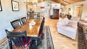 A spacious living room with a wooden ceiling, a dining area with a large table and chairs, and a kitchen in the background.