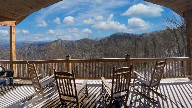 A wooden deck with four wooden chairs and a table, overlooking a mountainous landscape with a clear sky.