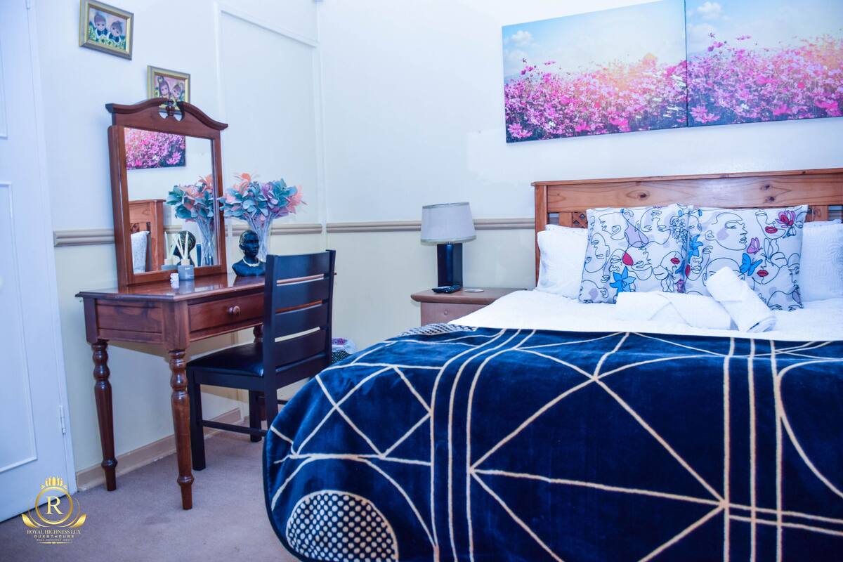 Een slaapkamer met een groot bed, een houten dressoir, een stoel, een nachtkastje, een lamp en een schilderij aan de muur.