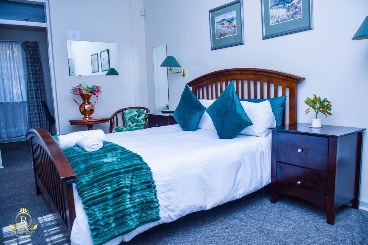 Een net opgemaakt bed met een groene deken, een houten hoofdbord en teal-kleurige kussens. Er staat een nachtkastje met een lamp en een potplant naast het bed.