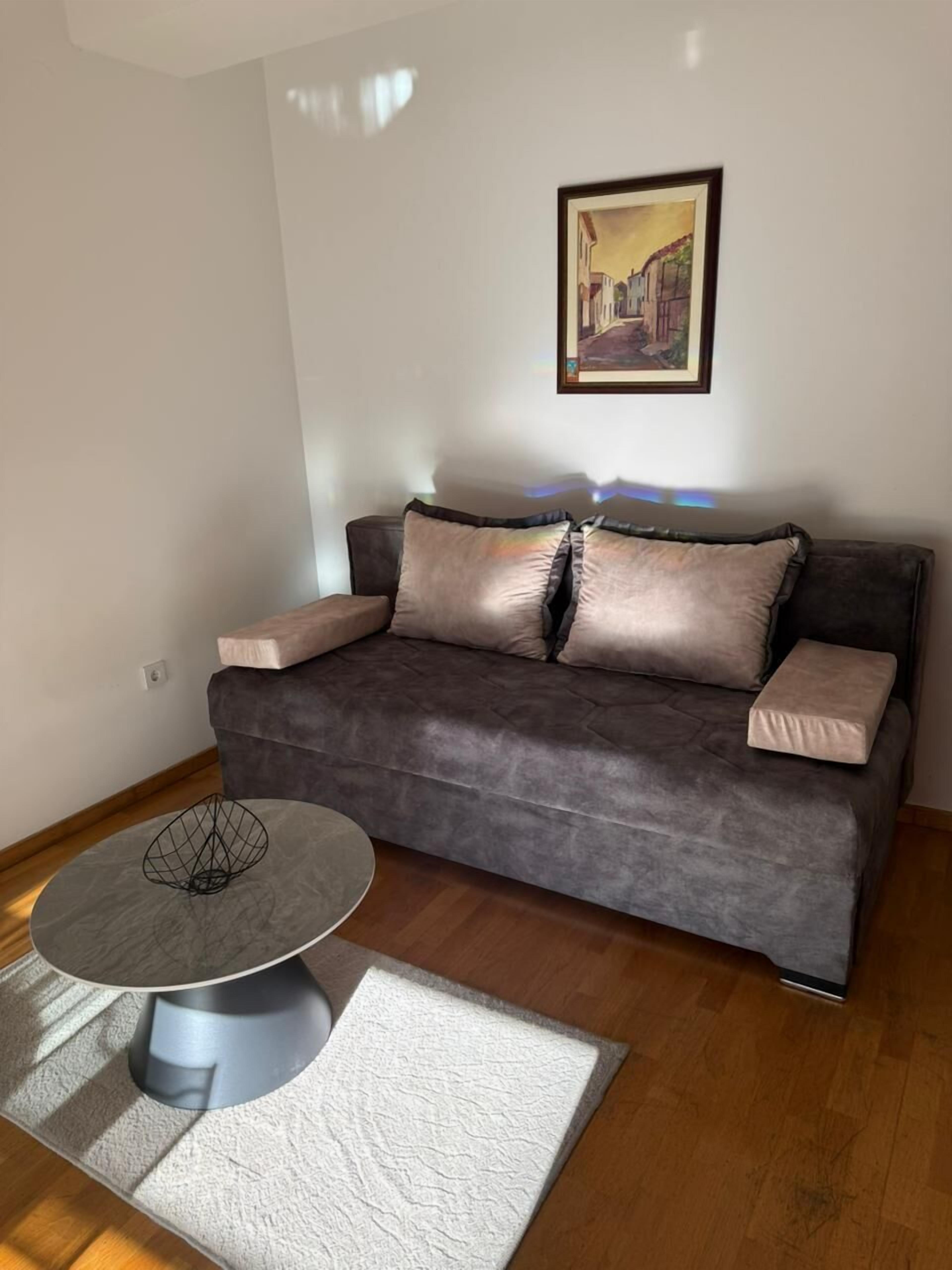 A living room with a dark grey sofa, two cushions, a framed picture on the wall, and a round table with a wireframe base.