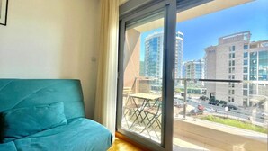 A modern room with a teal sofa, a large window with a view of city buildings and a balcony. - Comfort Apartments Aleksic (Budva)
