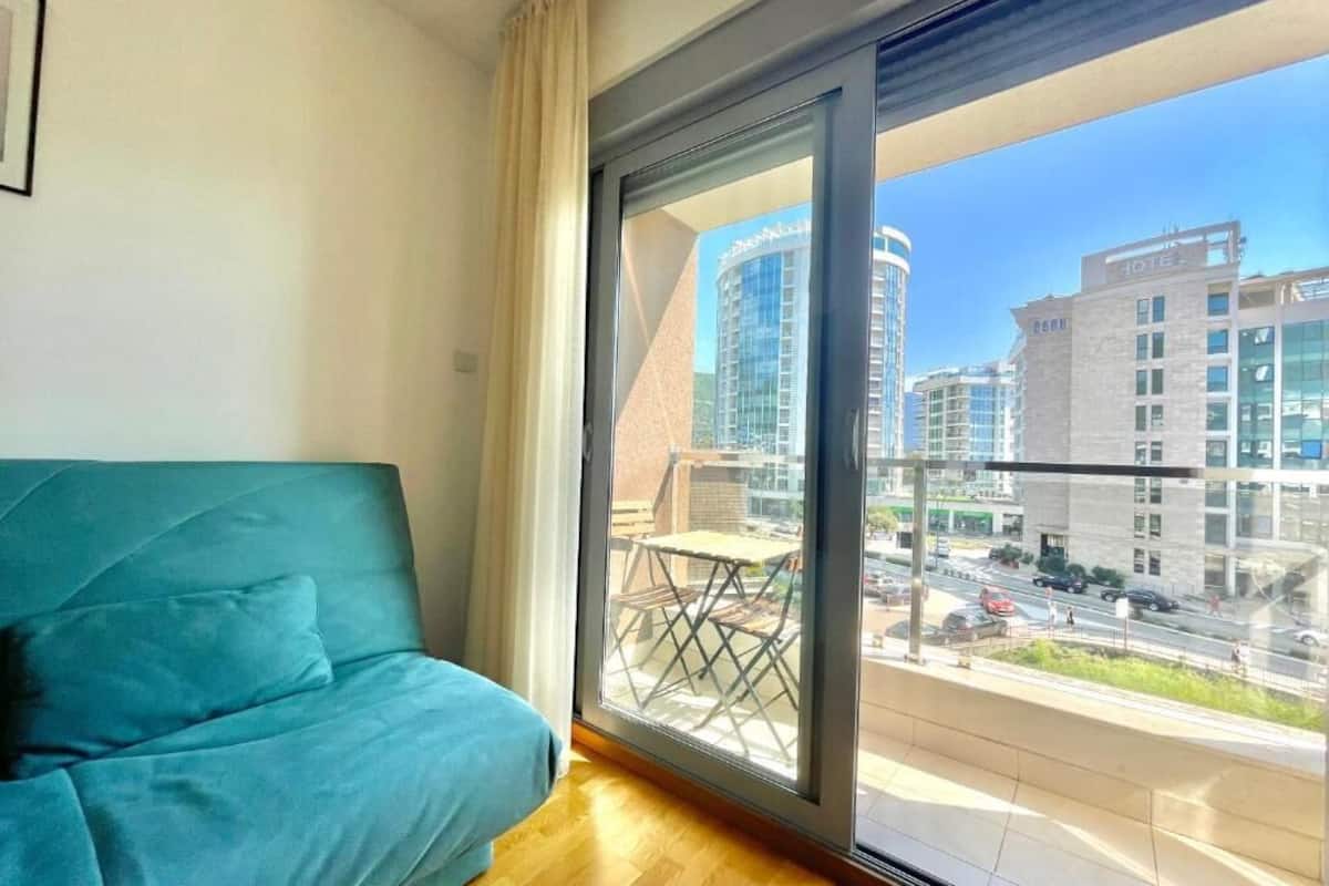 A modern room with a teal sofa, a large window with a view of city buildings and a balcony.